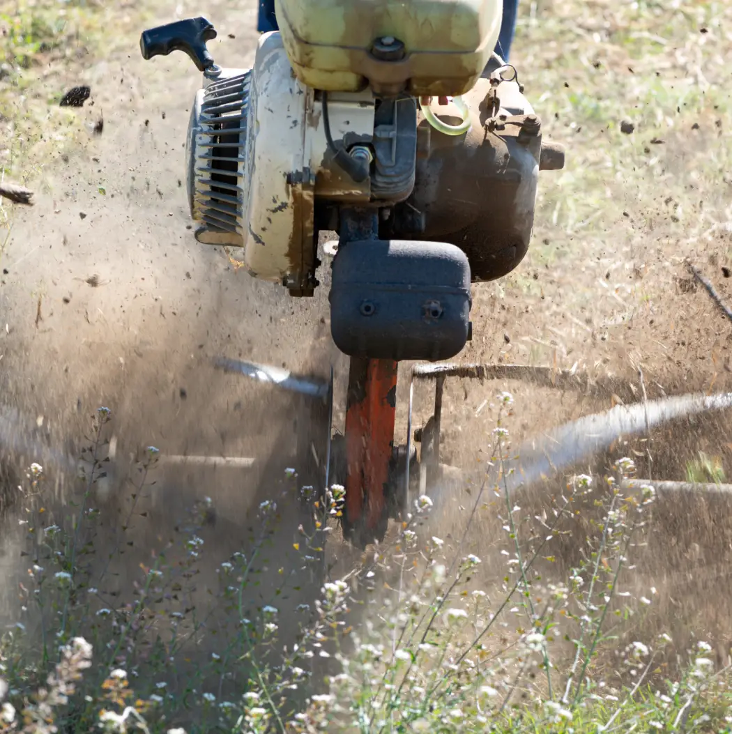 Layer-6241-1 A powerful tiller digs into the earth, creating a cloud of dust while aerating the soil. Surrounding greenery enhances the rugged nature of this agricultural scene, where machines meet nature in harmony.